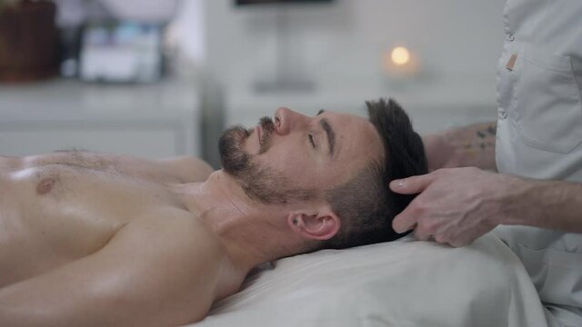 Side view of bearded brunette handsome man enjoying neck massage in spa indoors. Relaxed Caucasian male client lying on massage bed in beauty parlor as tattooed masseur rubbing skin in slow motion