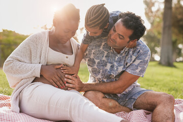 Multiracial family touching belly at picnic outdoor - African pregnant mother, caucasian father and...