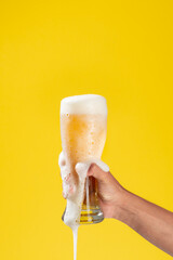 man's arm holds a glass beaker with beer inside, white foam and a solid yellow background, the foam falls through the glass and on the arm