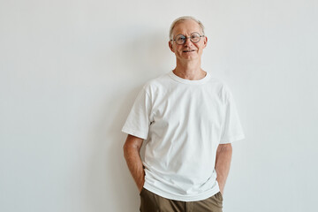 Minimal waist up portrait of smiling senior man wearing glasses while standing against white...