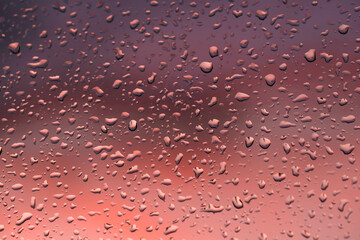 Szyba okienna pokryta od zewnątrz kroplami deszczu. Za oknem widać chmury zabarwione na czerwono światłem zachodzącego słońca. - obrazy, fototapety, plakaty