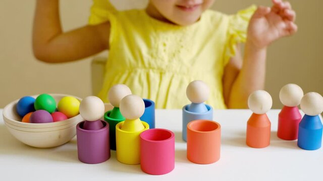 The girl learns colors by playing with wooden toy colored figures of a little man and puts them in cups of the appropriate color. After completing the task, the child cheerfully claps his hands