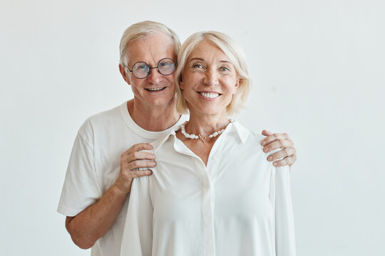 Minimal Waist Up Portrait Of Modern Senior Couple Embracing Against White Background And Looking At Camera