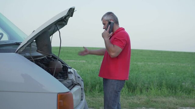 dissatisfied authentic man with beard and gray hair in red jersey near broken car minivan with raised hood indignantly talking on phone