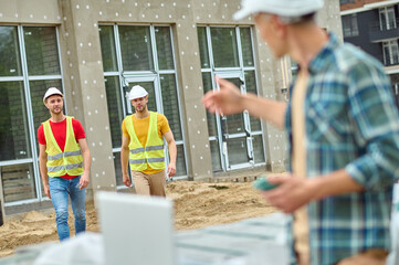 Serious construction workers in reflective waistcoats approaching their colleague