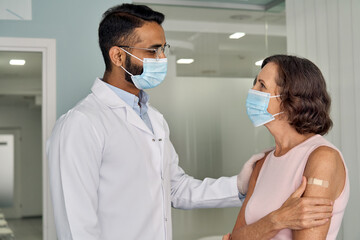 Happy young Indian doctor therapist in white coat wearing face mask supporting older senior female...