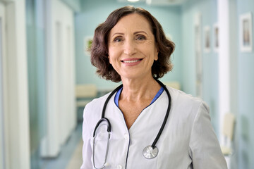 Portrait of smiling older senior female professional doctor physician pediatrician wearing white robe with stethoscope around neck standing in modern private clinic hospital, looking at camera. - obrazy, fototapety, plakaty