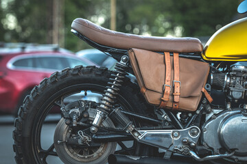 Vintage motorcycle detail with blurred car park on the background. Genuine leather bag for journeys. Beautiful, retro, shiny black motorbike with golden details and leather seat. Leather accessories - Powered by Adobe