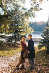Fototapeta premium A couple of beautiful lovers spend time outdoors on the fall day. Pretty woman in a red dress and modish hat and a handsome man in a raincoat posing at the city park on a sunny autumn day