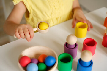 The girl puts colored balls in a wooden spoon and throws them into cups of the corresponding color. The child learns colors. A sorter toy for studying flowers by preschool children. A two-year-old kid