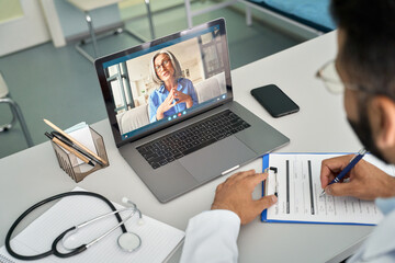 Over shoulder view of videocall chat with female older patient and indian male doctor in modern clinic hospital using laptop computer, consulting online remotely. Telemedicine healthcare concept.