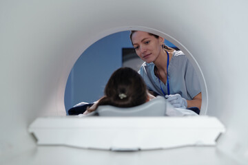 Young female clinician looking at little girl lying on table of mri scan machine before medical procedure - obrazy, fototapety, plakaty