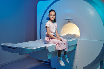 Smiling little patient sitting on long medical table before or after MRI scan procedure - obrazy, fototapety, plakaty