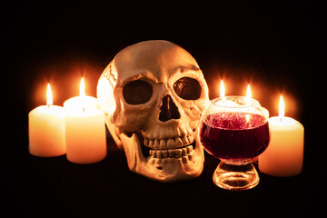 Human skull and glass of dark effervescent liquid among burning candles in the dark, scary still life, altar.