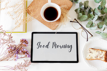Phrase"good morning" on a tablet, floral frame and breakfast composition on white background. Flat lay, top view. Slow morning concept.