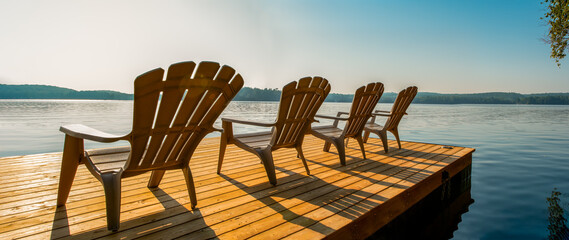 Row of Adirondack chairs -patio - deck chairs on wooden dock with sunset or sunrise -cottage life. Banner - panoramic - Powered by Adobe