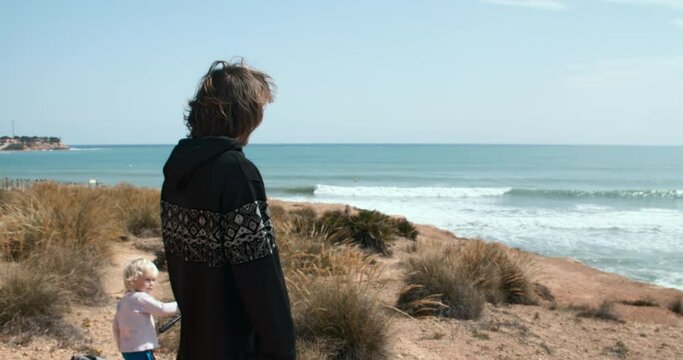 Young blogger man with phone in hand located on cliff expecting and showing waves in the sea. Little boy playing with tripod in background. Waves in mediterranean water at spring sunny day.