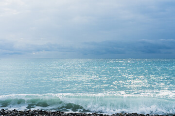 A wave of turquoise sea shining in the sun runs over black pebbles. The sea on the horizon merges with the blue sky.