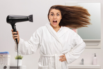 Surprised young woman in a bathrobe blow drying hair - obrazy, fototapety, plakaty