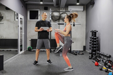 Obraz na płótnie Canvas Sports training performance and progress measurement. A woman in sportswear does a high skip indoor gym while the trainer observes her training and enters data into a digital tablet