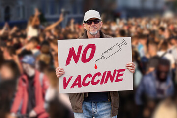 Displeased man with cap, blue jeans and sunglasses holding a NO COVID vaccine sign with crowd of people in background. Supporting anti-vaccination movement. Anti-vaxxer activist.  - obrazy, fototapety, plakaty