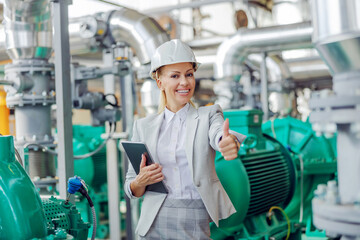 Middle-aged successful blond female supervisor in suit with protective helmet on head standing in power plant with tablet in hands and showing thumbs up.