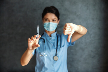 Doctor with face mask holding syringe with vaccine. She is showing thumbs down. It's not safe to get covid-19 vaccine. Selective focus on hands.