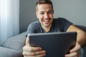 Online chat and internet messaging, phone call. A man with a beautiful smile discusses with friends and colleagues. He has white wireless headphones and uses a gray tablet that he holds in his hands