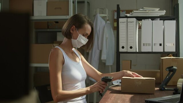 Young woman in protective mask working in storage, scanning barcode of parcel, typing on laptop keyboard. Woman sitting at workplace in storage area, warehousing concept. Small business during covid.
