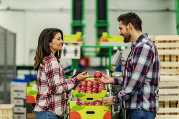 A couple in plaid clothes chooses apple from the crates in a production warehouse and evaluates a quality apple that is recorded on a tablet Man handed the apple to the girls and they made eye contact
