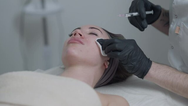 Young woman lying in beauty parlor as professional beautician injecting botox filler in face with syringe. Caucasian beautiful slim female client in spa and expert with syringe and needle. Slow motion