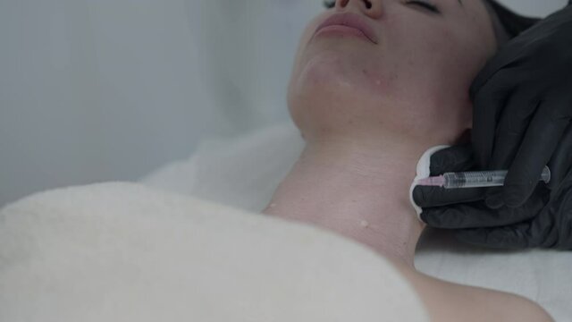 Close-up beauty injection with syringe in slim female Caucasian neck. Beautician in black gloves injecting botox filler for beautiful woman lying in spa parlor indoors. Slow motion
