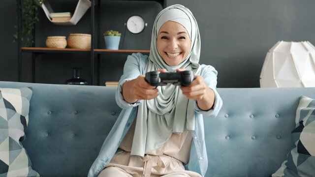 Portrait of cheerful muslim woman in hijab enjoying video game having fun looking at camera indoors at home. Leisure time and entertainment concept.