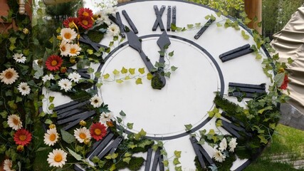 Large wall clock on the field in blooming flowers, showing the time 11:55, 12:00. time management concept, daylight saving time. Spring or wedding. Picnic for womens day