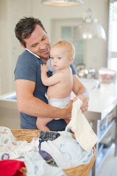 Father Holding Baby And Folding Laundry While Talking On Cell Phone