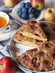 Laid table with freshly baked summer round pie with apples and plums in a white ceramic tin. Sliced piece of cake and ingredients on a white wooden table. Food background.