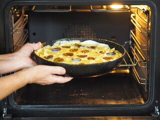 An elderly woman is hands put a pan of apple and plum dough into the kitchen oven. Woman is hobby. household chores. Preparation for a holiday or meeting guests.