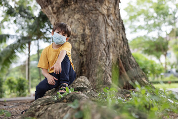 Child wearing face mask sitting under big tree and looking at camera.Pollution concept.