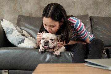 A beautiful, cute casual Asian woman is happily fun playing, teasing, and hug adorable dog (French Bulldog) on grey sofa in living room of house. Pet is her family and friends in lifestyle.