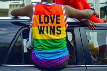 A young man sitting in a car window wears a T-shirt with the LGBTQ flag and it has LOVE WINS written on it.
