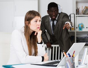 Frustrated woman sitting at office desk with disgruntled boss behind