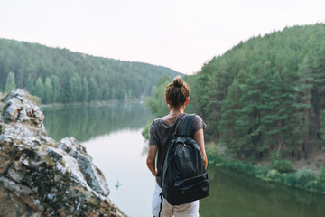 Fototapeta na wymiar Young slim woman in casual clothes with backpack looks at the beautiful view of mountains and calm river, local travel