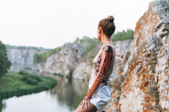 Young slim woman in casual clothes looks at the beautiful view of mountains and calm river, local travel