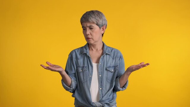 Confused puzzled upset middle aged Asian woman standing isolated over yellow background