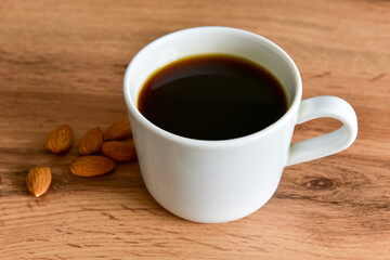 White cup of strong black coffee with almonds lying nearby on a wooden surface close-up
