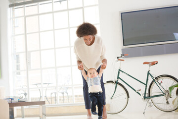 Mother helping baby boy walk in living room