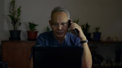 Asian senior businessman talking on mobile phone with company coworker while watching business data from laptop monitor. Elderly male professional working on the desk at home. Late working.  - Powered by Adobe