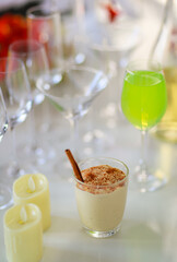 Delicious tasty glass of creamy spicy eggnog with gingerbread on decoration Thanksgiving Christmas eve party table full of empty various tall wine glasses green apple punch white chocolate candles