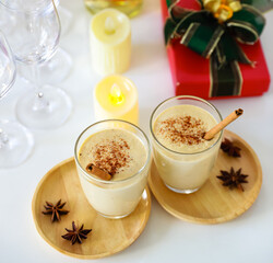 Top view shot of two sweet creamy alcoholic eggnog drinks with gingerbread on wooden coaster with anise on white clothing table full of empty tall wine glasses liquor bottles and present gift boxes
