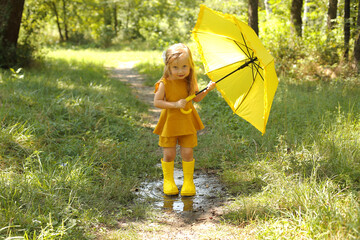 a beautiful blonde girl in a linen suit in yellow boots with a yellow umbrella stands in the rain in the park
